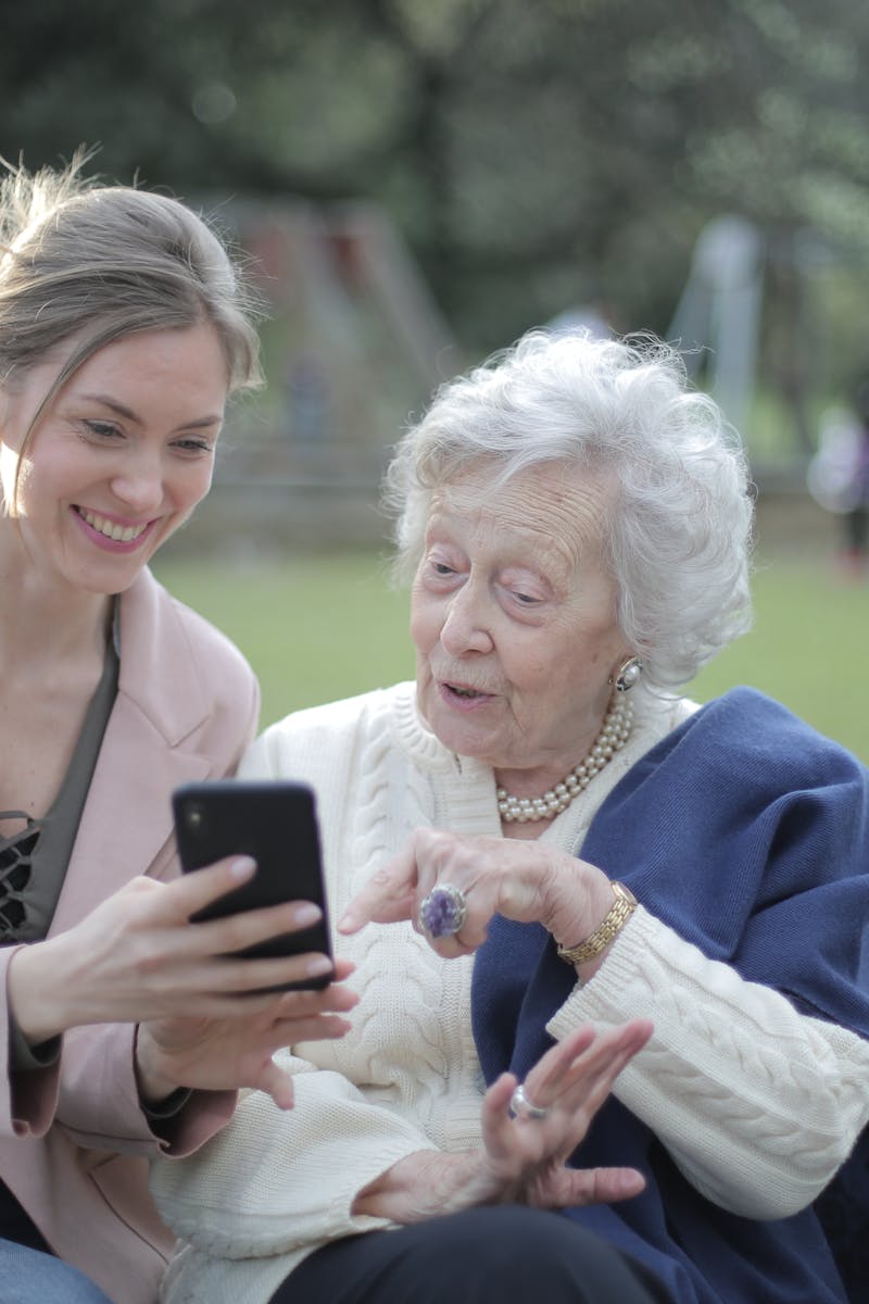 Delighted female relatives sitting together on wooden bench in park and browsing mobile phone while learning using