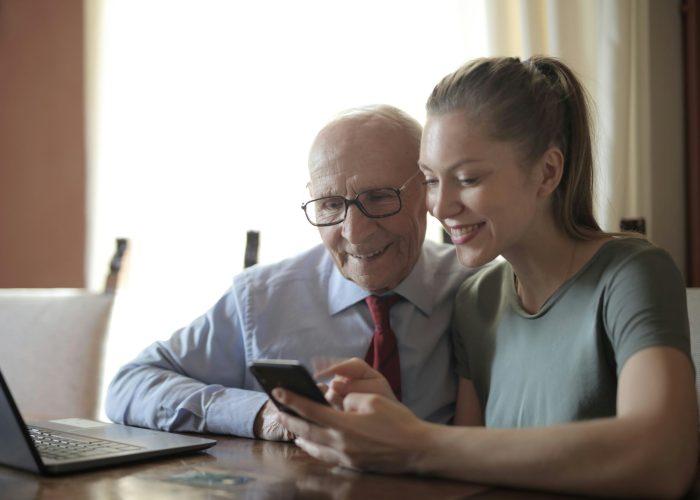 pexels-photo-3823542-3823542 Smiling young woman in casual clothes showing smartphone to interested senior grandfather in formal shirt and eyeglasses while sitting at table near laptop